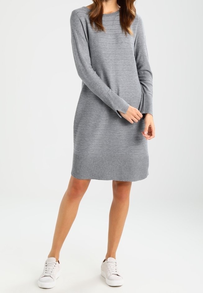 Femme portant une robe-pull grise à manches longues et longueur genou, ainsi que des baskets blanches, debout avec une main touchant l'autre poignet.