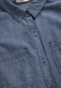 Tummansininen denimpaitapusero, jossa on klassinen kaulus, kaksi rintataskua ja pyöreät napit. Tekstuuriainetta, jossa on pehmeä pinta.