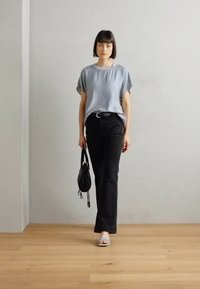 Haut gris à manches courtes, jeans noirs à coupe droite et sandales bleu clair. Un petit sac à main noir avec une sangle est porté. Sol en bois et fond uni.