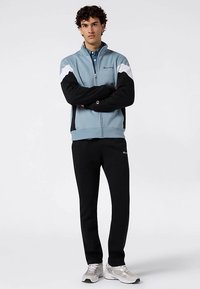 Giacca con zip azzurro chiaro con accenti neri e bianchi, abbinata a pantaloni sportivi neri. Il tessuto sembra morbido, adatto per un abbigliamento casual.