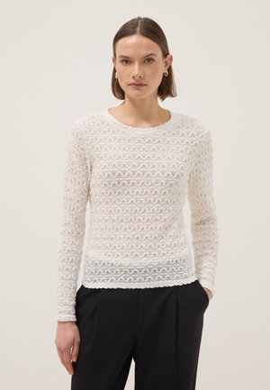 Mujer con cabello corto y castaño, que lleva pendientes de aro dorados, una blusa blanca de manga larga y textura, y pantalones negros, de pie con una mano en el bolsillo.