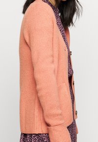 Růžový pletený kardigan s žebrovanou strukturou, s knoflíkovým zapínáním a bočními kapsami, nošený přes vzorované šaty.