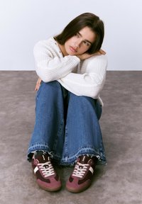 Joven mujer con suéter blanco, jeans anchos y zapatillas burdeos está sentada en el suelo, apoyando la cabeza en los brazos doblados sobre las rodillas, mirando hacia un lado.