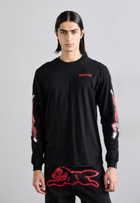 Maglietta nera a maniche lunghe con accenti grafici rossi sulle maniche, caratterizzata da un logo sul petto. Abbinata a jeans scuri con un design stampato in rosso.