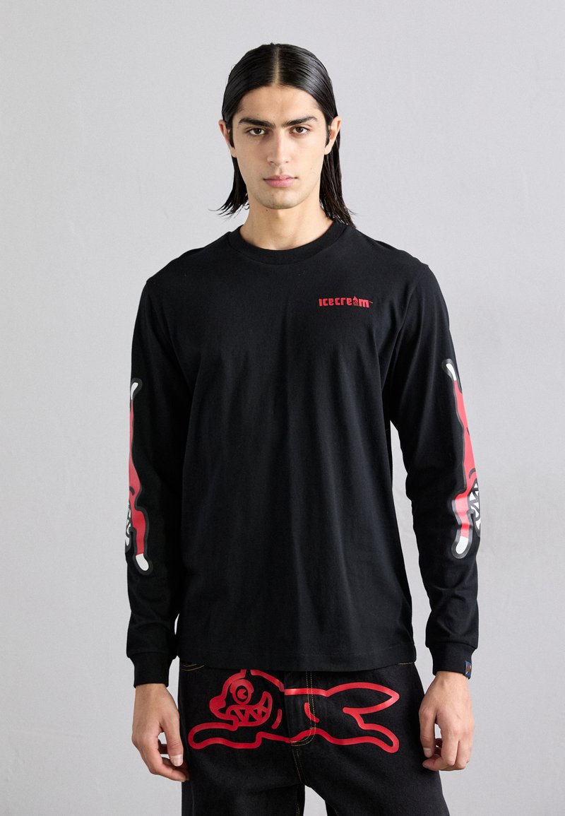 Maglietta nera a maniche lunghe con accenti grafici rossi sulle maniche, caratterizzata da un logo sul petto. Abbinata a jeans scuri con un design stampato in rosso.