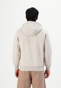 Man in een lichtgrijze capuchontrui en beige broek, staand met zijn rug naar de camera tegen een effen witte achtergrond.