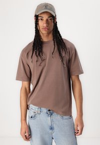 Brown cotton t-shirt with short sleeves, relaxed fit, and distressed details, paired with faded blue denim jeans. Accent cap in a light color.