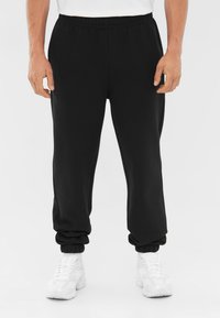 Zwarte sportbroek met een elastische tailleband en manchetten, gemaakt van gladde stof, gecombineerd met witte sportschoenen.