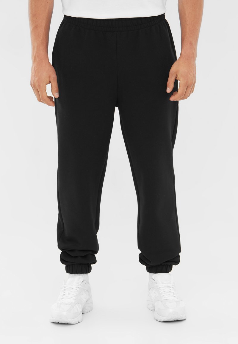 Zwarte sportbroek met een elastische tailleband en manchetten, gemaakt van gladde stof, gecombineerd met witte sportschoenen.