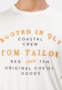 Λευκό T-shirt με πορτοκαλί και μαύρο κείμενο που γράφει "Rooted in Qly Coastal Crew Tom Tailor Reg 1962 TRM Original Custom Goods"