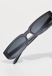 Lunettes de soleil rectangulaires noires avec des verres sombres, monture en plastique épaisse et texture lisse. Les branches sont légèrement effilées avec une finition brillante.