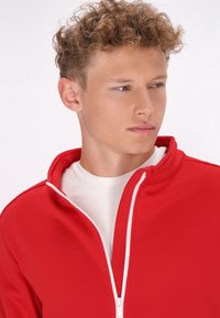 Rode ritsjack gemaakt van zachte stof, met een hoge kraag en een witte rits. Draagt over een wit shirt, met een close-up van de details.