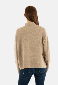 Jersey beige de punto con cuello alto y corte holgado, con dobladillo acanalado. Combinado con jeans oscuros, visto desde la espalda.