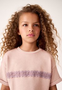 Šviesiai rožinis džemperis, pagamintas iš minkštos medžiagos, puoštas dekoratyvine alyvinės spalvos nėrinių juosta su tekstu per krūtinę. Trumpomis rankovėmis ir apvalia iškirpte.