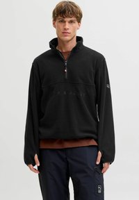 Młody mężczyzna ubrany w czarną polarową bluzę z zamkiem błyskawicznym z napisem "Jack & Jones", brązową koszulę pod spodem oraz ciemne spodnie cargo z naszywką Tech Fusion.