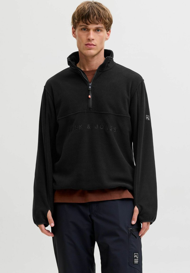 Młody mężczyzna ubrany w czarną polarową bluzę z zamkiem błyskawicznym z napisem "Jack & Jones", brązową koszulę pod spodem oraz ciemne spodnie cargo z naszywką Tech Fusion.
