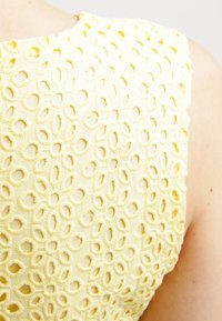 Un tejido amarillo texturizado cuenta con un diseño de ojales florales con recortes circulares. La prenda tiene un escote redondeado y es ligera.