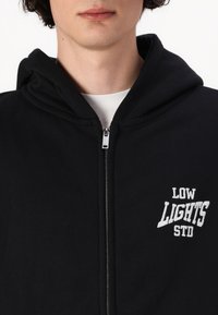 Svart zip-up hettegenser laget av mykt stoff, med en hvit brodert logo "LOW LIGHTS STD" på venstre side. Hette er festet.