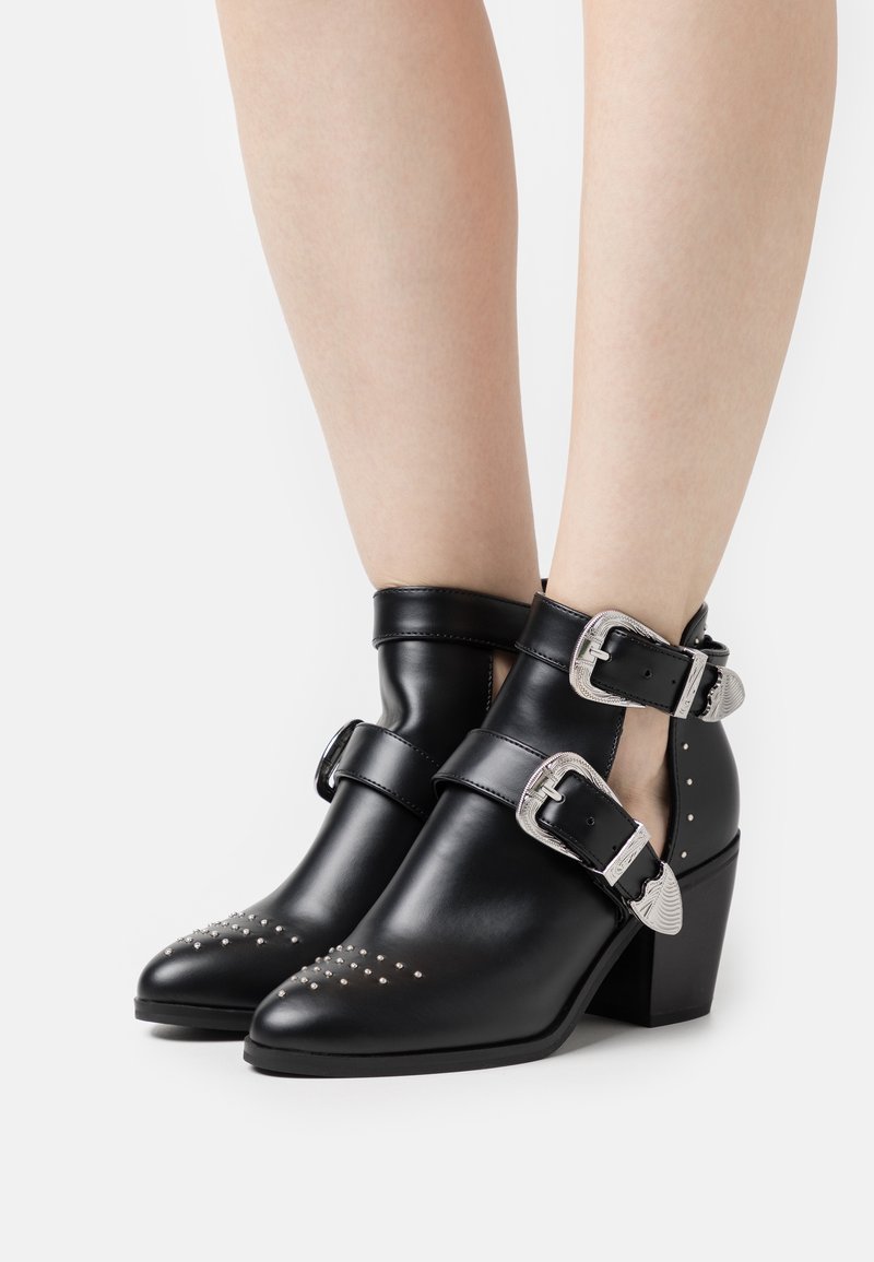 Personne portant des bottines noires à boucles argentées, bouts pointus, talons larges et bas, et petits clous métalliques sur les bouts, debout sur fond blanc.