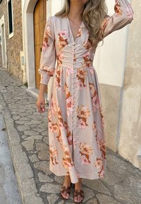 Florale maxi-jurk in lichtroze met oranje en roze rozenpatronen. Beschikt over een V-hals, lange puffmouwen en knoopdetails aan de voorkant.