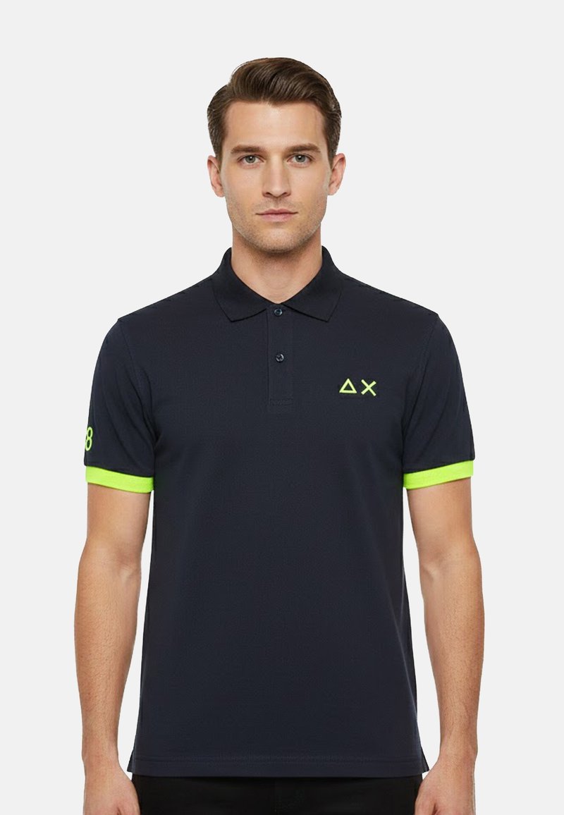 Homme portant un polo noir avec un liseré vert néon sur les manches et des symboles géométriques sur la poitrine et la manche, faisant face à l'avant.