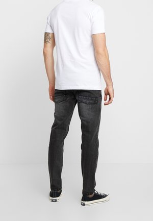 Mann trägt weißes T-Shirt, dunkelgraue Jeans und schwarze niedrige Sneakers und steht mit dem Rücken zu vor einem schlichten hellen Hintergrund.