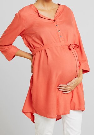 Femme enceinte en tunique de maternité corail à manches trois-quarts et pantalon blanc, main posée sur le ventre et le bas du dos.