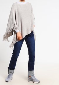 Poncho grigio chiaro in tessuto testurizzato con dettagli in frange, abbinato a jeans blu scuro. Il modello indossa sneaker grigie.