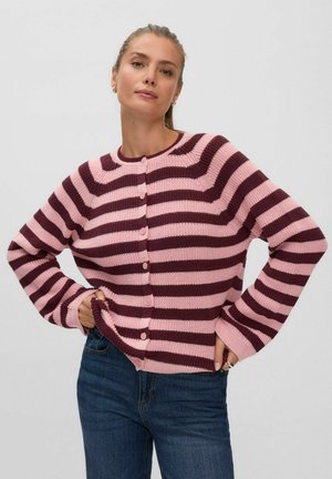 Femme portant un cardigan boutonné rayé rose et rouge foncé à manches longues et un jean bleu, debout les mains sur les hanches.