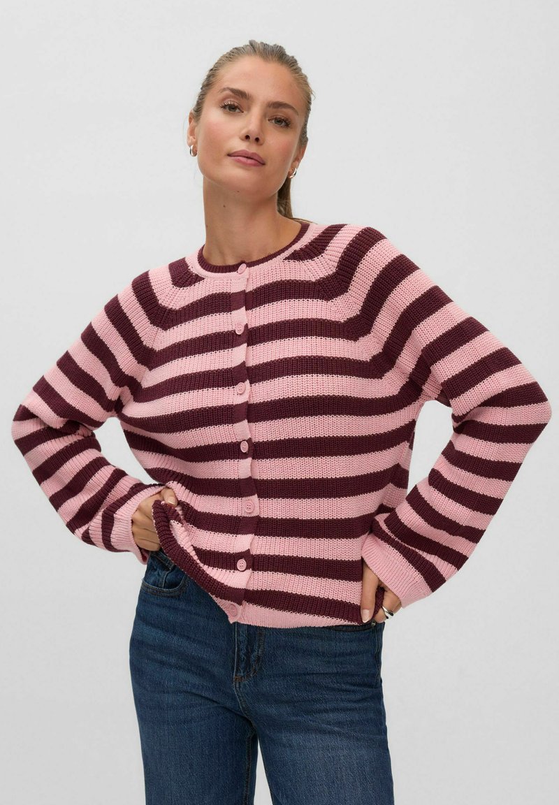 Femme portant un cardigan boutonné rayé rose et rouge foncé à manches longues et un jean bleu, debout les mains sur les hanches.