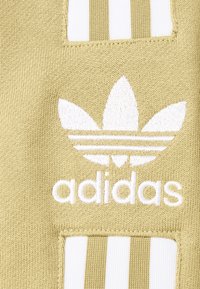Olivgrön tyg med vit adidas-logotyp och tre ränder, med en texturerad stickad bakgrund och ett patchdesign.