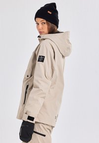Beige waterdichte jas met capuchon, ritszakken en klittenbandmanchetten. Zwarte beanie met een leren label en zwarte wanten met tekstdetail.