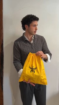 Jonge man met krullend haar die een grijze trui draagt, houdt een felgele trekkoordtas met een zwart embleem vast en kijkt naar rechts.