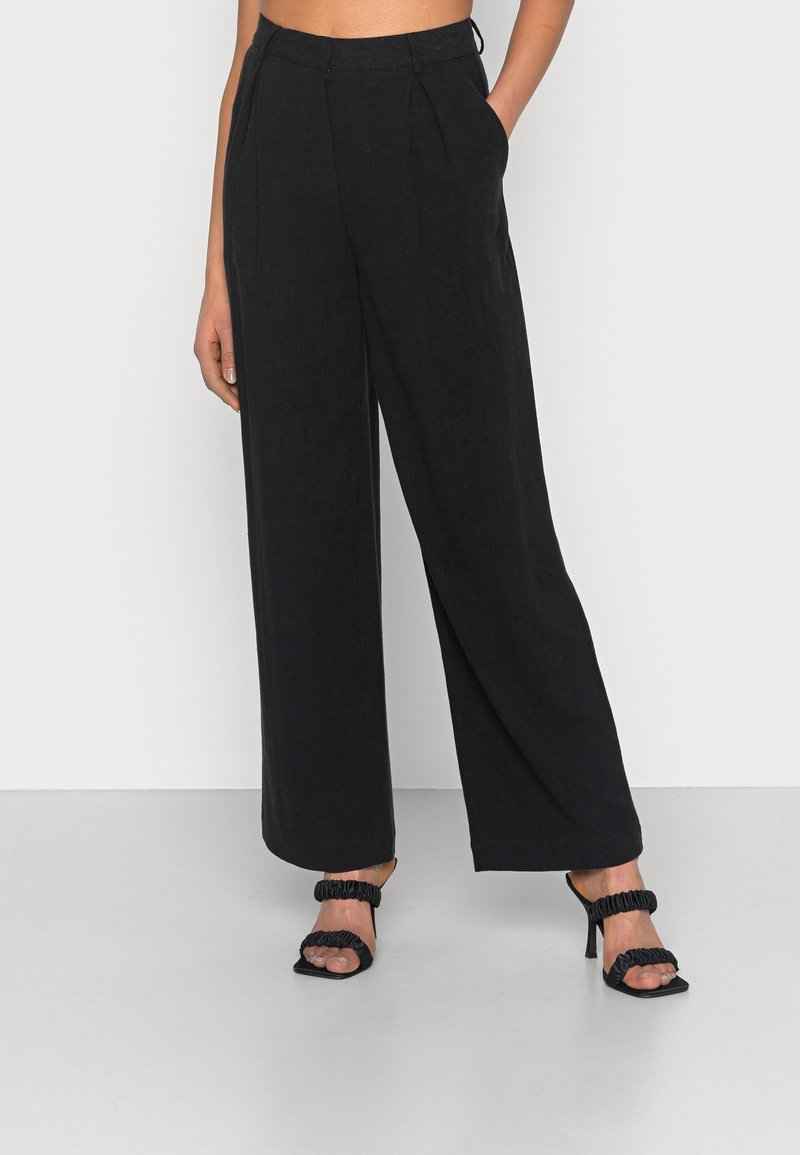Femme portant un pantalon noir taille haute à jambes larges, mains dans les poches, associé à des sandales noires à talons ouverts sur un fond uni.