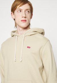 Lys beige hettegenser med frontlomme, snøring på hetten og en liten rød Levi's-logo på brystet. Myk stoffkvalitet og avslappet passform.