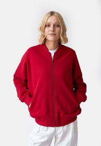 Czerwony sweatshirt na zamek błyskawiczny z okrągłym dekoltem, luźny krój, boczne kieszenie i ściągacze na mankietach, w połączeniu z białymi spodniami. Gładka faktura materiału.