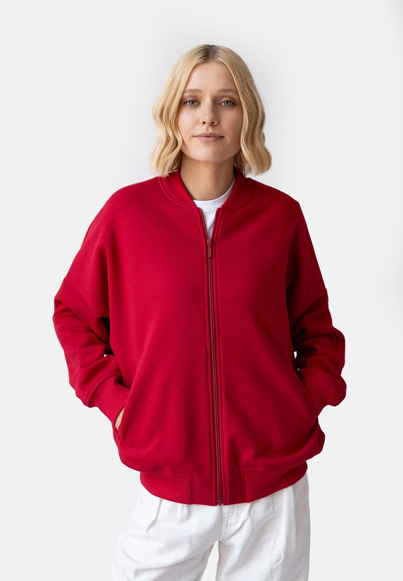 Czerwony sweatshirt na zamek błyskawiczny z okrągłym dekoltem, luźny krój, boczne kieszenie i ściągacze na mankietach, w połączeniu z białymi spodniami. Gładka faktura materiału.