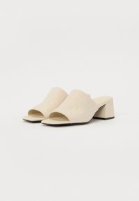 Mules beige à talon bloc avec un bout ouvert. Tige texturée présentant un motif subtil. Semelle plate avec un accent noir à la base du talon.