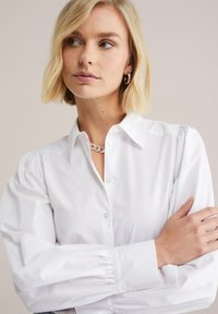 White button-up shirt with a collar, featuring puffed sleeves and gathered cuffs. Smooth texture with a classic design and minimal detailing.