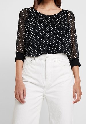 Femme portant un chemisier noir transparent à pois blancs avec des manches trois-quarts, associé à un pantalon blanc taille haute.