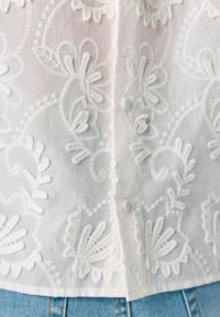 Blouse blanche brodée avec des motifs floraux et des coutures en relief, fermetures à bouton et une structure en tissu léger.
