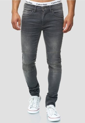 Homme portant un jean noir slim délavé avec des détails côtelés aux genoux et des baskets blanches sur un fond uni.