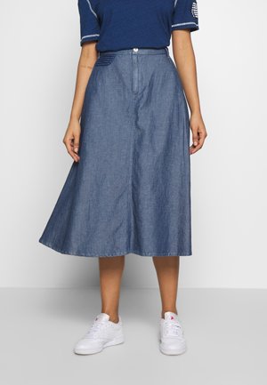 Kvinde iført en blå midilæng denimnederdel, marineblå t-shirt og hvide sneakers, stående foran en ensfarvet lys baggrund.