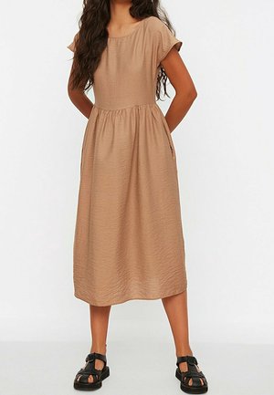 Vrouw met lang haar, gekleed in een beige midi-jurk met korte mouwen en zwarte gesloten sandalen, staand met handen achter de rug.