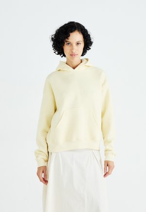 Light yellow hooded sweatshirt with a front pocket, relaxed fit, and ribbed cuffs. Smooth fabric with a simple design. Underneath is a white skirt.