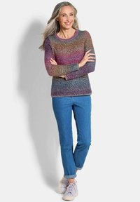Femme aux longs cheveux gris souriante, portant un pull en laine multicolore, un jean bleu retroussé et des baskets lilas claires, les bras croisés.
