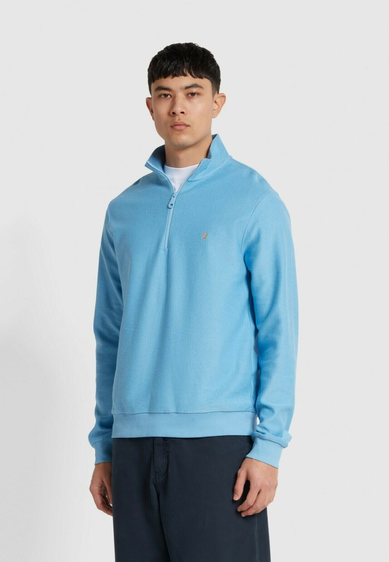Lichtblauwe pullover met rits in een gebreide textuur. Heeft een ontspannen pasvorm, geribde manchetten en een klein logo op de borst.