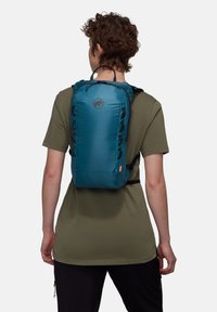 Άτομο που φοράει ένα μικρό σακίδιο πλάτης σε πετρόλ χρώμα με λογότυπο ελέφαντα, πράσινο ελιάς πουκάμισο και μαύρο παντελόνι, με την πλάτη γυρισμένη πάνω σε λευκό φόντο.