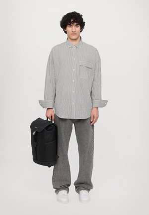 Camicia a righe con taschino sul petto, pantaloni grigi a gamba larga e zaino nero con chiusura a rotolo. Scarpe bianche completano l'outfit.