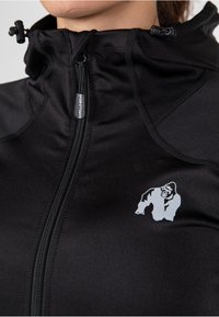 Zwarte sportjack met hoge kraag, ritssluiting en een grijze gorilla-logo op de linkerborst. Soepele, rekbare stof met zichtbare stiksel.